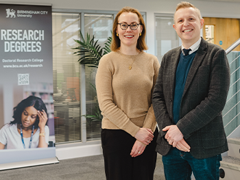 BCU's Jenny Harding and Professor Oliver Carter pictured next to a research degrees banner on securing a Doctoral Landscape Award from the AHRC