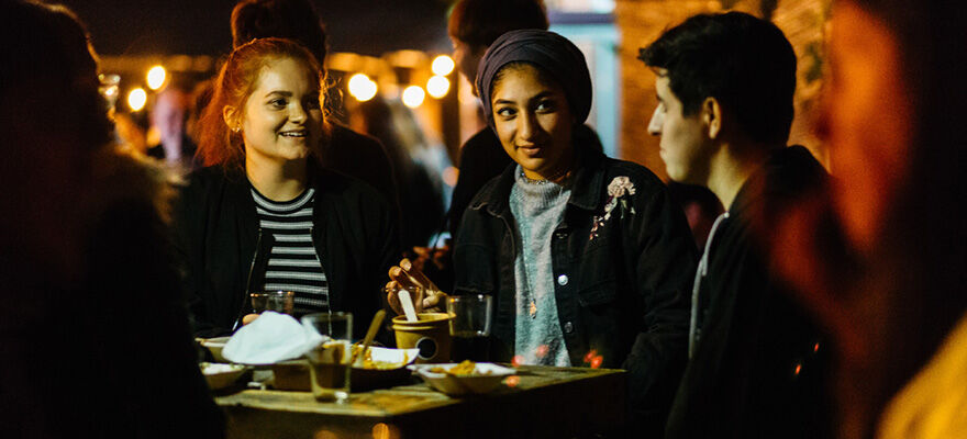 Three students sitting together at Digbeth Dining Club. There's warm festoon lighting in the background and they're smiling listening to one person speaking