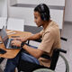 A young man in a brown t-shirt is working on a laptop at a desk while sitting in a wheelchair