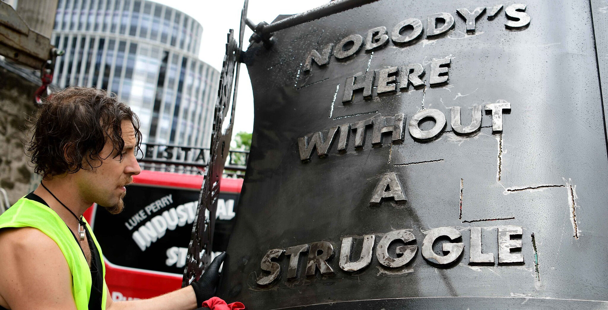 Image of artist Luke Perry in high-vis vest adding the final touches to his steel art crest which shows the words 'Nobody's here without a struggle'