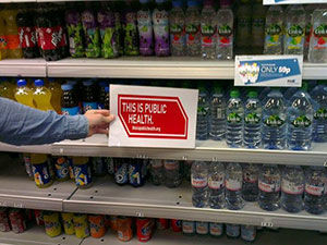 'This is Public Health' sign being held up in a shop with drinks in the background