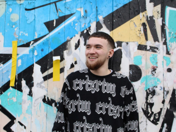Charlie Adams, a young white man with dark hair is looking off camera and smiling. He's wearing a black jumper with white graphic design and is standing in front of a brightly coloured graffitied wall