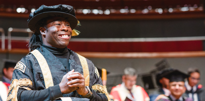 Chancellor Ade Adepitan at graduation. He is smiling at the crowd and wearing graduation robes.