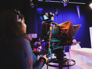 A student is standing behind the camera looking in the view finder. She is wearing headphones and a wavy top. Her camera is pointed at a TV studio set with white furniture and a purple background