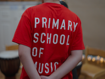 A child is wearing a red t-shirt with white text that reads 'Primary School of Music'