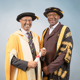 Tom Ilube CBE (left) and Chancellor Sir Lenny Henry are standing in front of a grey background. They are wear graduation robes and smiling at the camera