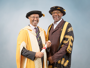 Tom Ilube CBE (left) and Chancellor Sir Lenny Henry are standing in front of a grey background. They are wear graduation robes and smiling at the camera